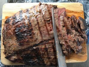 Grilled flank steak being sliced on cutting board for Flank Steak Salad Recipe with halved grape tomatoes, and creamy basil yogurt dressing.