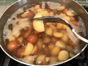 Fork in a cooked potato over a pot of boiling potatoes for red potatoes for Red Potato Salad Recipe with Edamame and Harissa. It's creamy, crunchy, meaty, tangy, peppery, salty, and oh, it's UNBELIEVABLE!