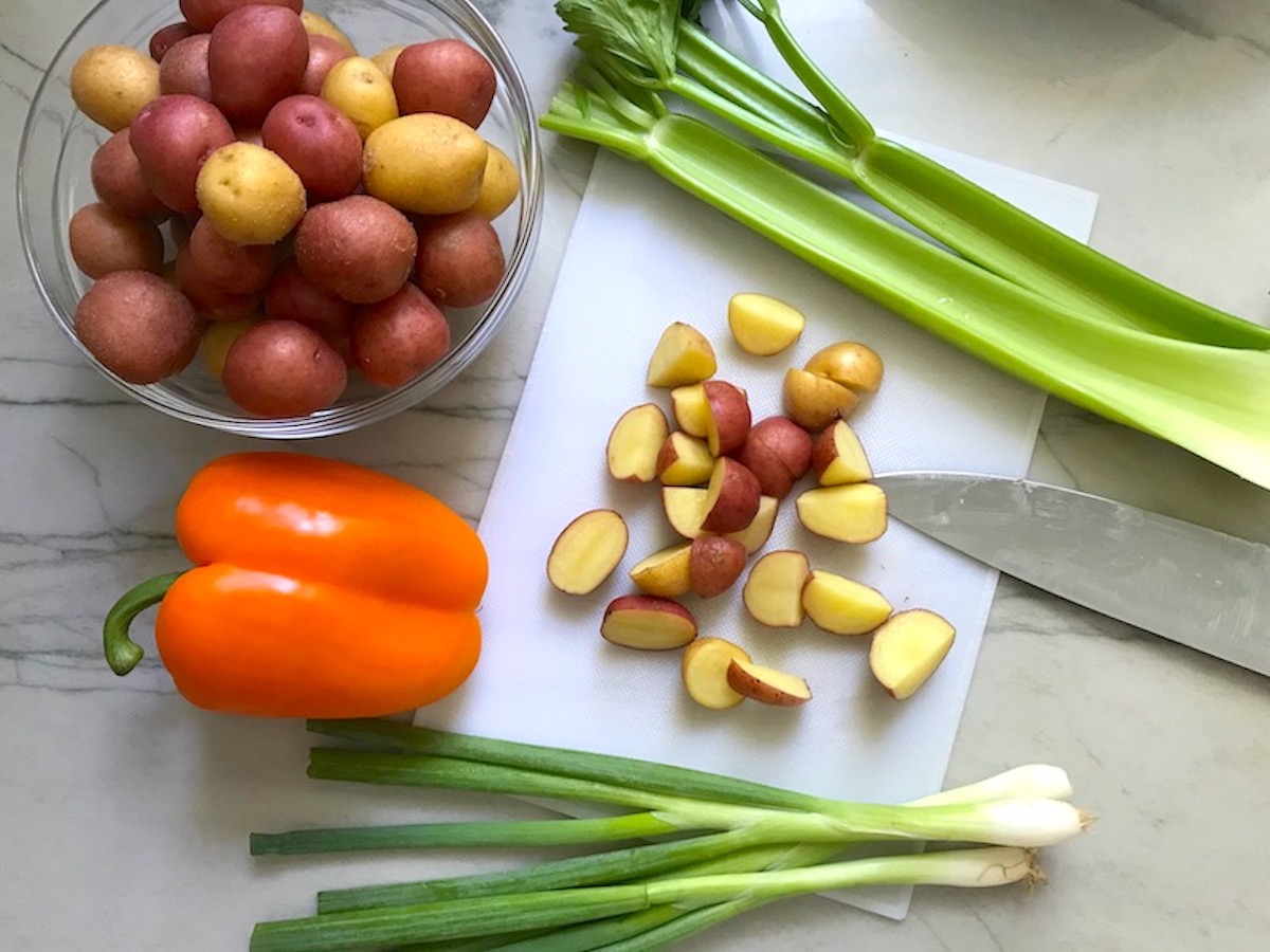 Baby creamer potatoes in a bowl and on a cutting board cut in quarters for Red Skin Potato Salad Recipe. Also a whole orange bell pepper, celery stalks, and whole scallions on counter.