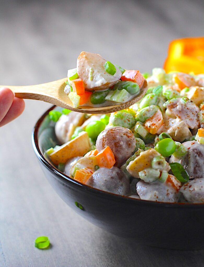 Bamboo spoon holding scoop of Red Skin Potato Salad Recipe with Edamame and Harissa in a bowl on table. It's creamy, crunchy, meaty, and delicious.
