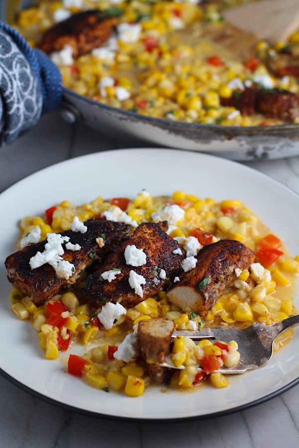 Creamy Corn and Blackened Chicken with feta and cilantro on top on a plate with fork holding a bite of chicken..