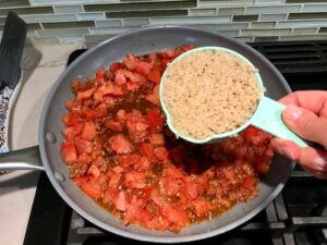 1 cup measuring cup adding cooked quinoa to chorizo and tomatoes in pan for Stuffed Tomatoes with chorizo, quinoa, parmesan cheese, chives, garlic, and oregano.