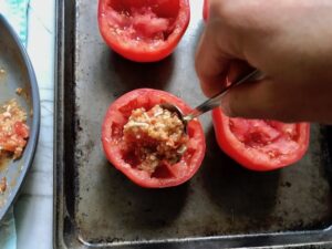 Hand spooning filling into a tomato on sheet pan for Stuffed Tomatoes with chorizo, quinoa, parmesan cheese, chives, garlic, and oregano.