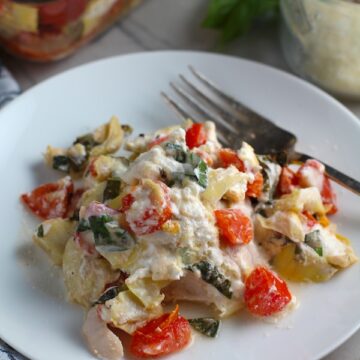 Chicken Artichoke Bake with Tomatoes and Goat Cheese on a plate with fork and casserole in back. It's a creamy, tangy, sweet, savory, mouth watering easy dinner that you can prep ahead!