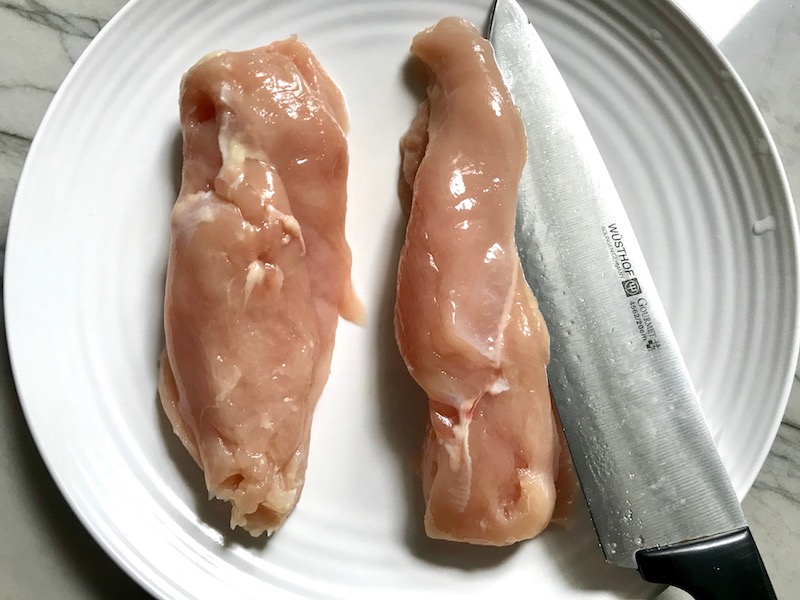 Raw chicken breasts being sliced from the side with knife for Chicken Parmesan Pasta Bake. It cooks entirely in one casserole dish, including the pasta! Shell pasta in flavorful, tangy tomato sauce is topped with zucchini, chicken, mozzarella, parm, and crunchy breadcrumb topping.
