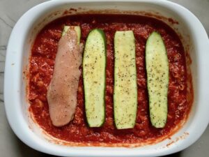 Zucchini and chicken being layered on pasta in casserole dish for Chicken Parmesan Pasta Bake. It cooks entirely in one casserole dish, including the pasta! Shell pasta in flavorful, tangy tomato sauce is topped with zucchini, chicken, mozzarella, parm, and crunchy breadcrumb topping.