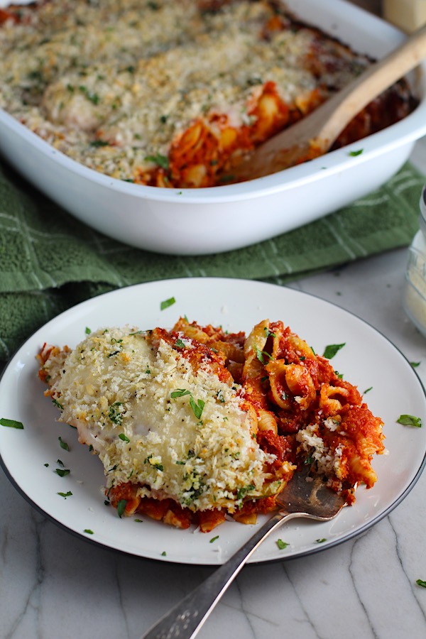 Chicken Parmesan Pasta Bake on a plate with casserole in back. It cooks entirely in one casserole dish, including the pasta! Shell pasta in flavorful, tangy tomato sauce is topped with zucchini, chicken, mozzarella, parm, and crunchy breadcrumb topping.