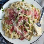 Easy Fettuccine Alfredo with Prosciutto on a plate with fork scooping pasta. It's a creamy but lighter recipe without the heavy cream or butter, but still delicious!