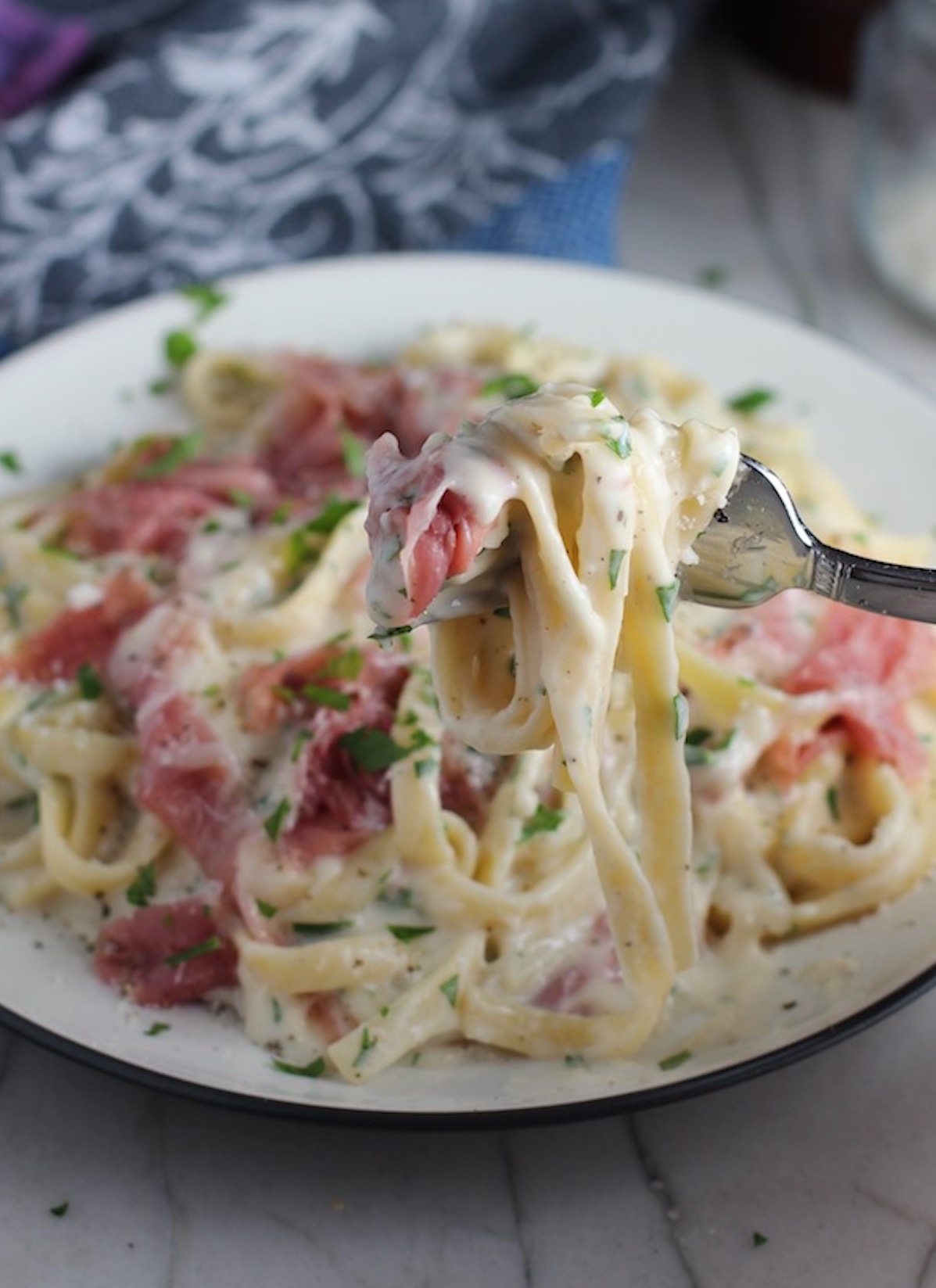 Fork with bite of Fettuccine Lower Fat Alfredo Sauce Recipe with Prosciutto with plate of the pasta below. It's a creamy but lighter recipe without the heavy cream or butter, but still delicious!