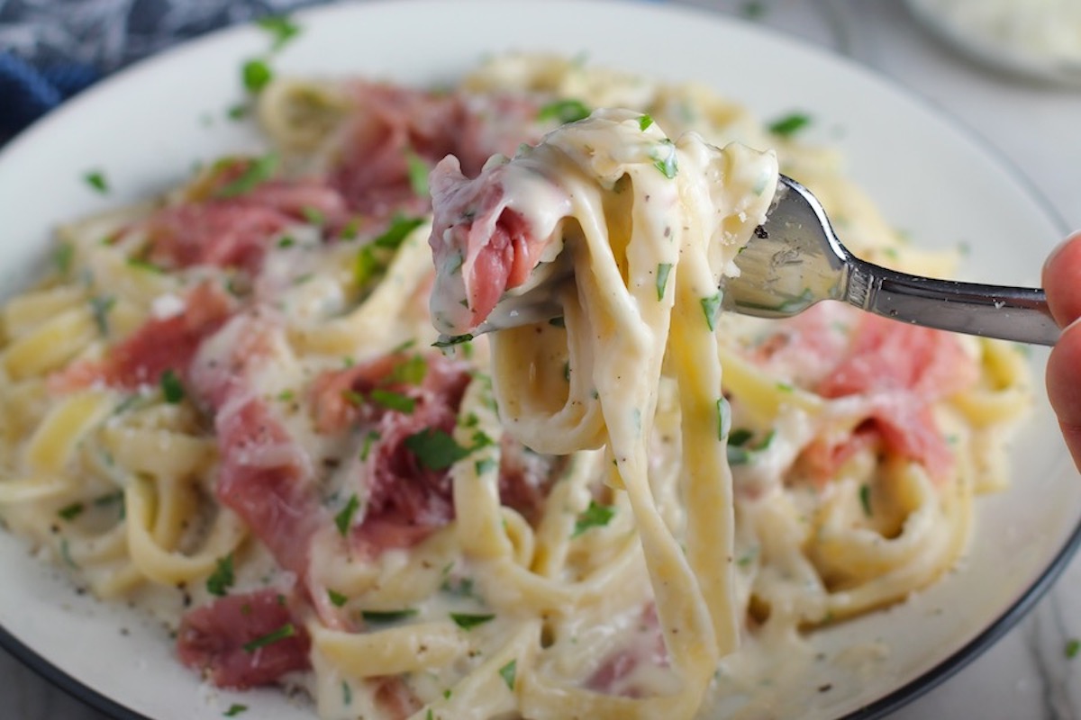 Fork with bite of Fettuccine Lower Fat Alfredo Sauce Recipe with Prosciutto with plate of the pasta below. It's a creamy but lighter recipe without the heavy cream or butter, but still delicious!