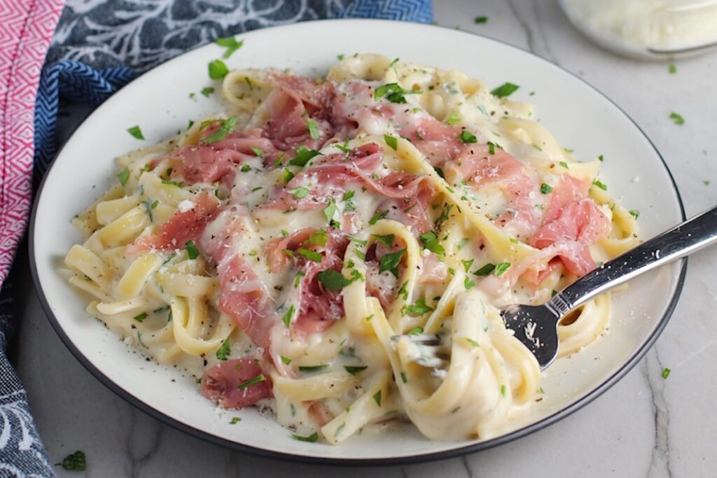 Fettuccine Lower Fat Alfredo Sauce Recipe with Prosciutto on a plate with fork scooping pasta. It's a creamy but lighter recipe without the heavy cream or butter, but still delicious!