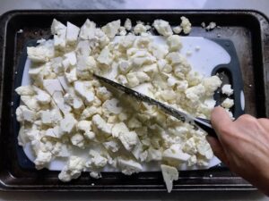 Knife cutting cauliflower on cutting board in sheet pan for Roasted Garlic Cauliflower Mash.