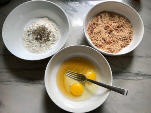 3 bowls on counter for Oven Fried Chicken Breast. One bowl has flour, another has honey and eggs, and the last has breadcrumbs. The chicken is coated with Bacon Crumbs and Honey and it's healthier because it's baked, not fried, but still gives you all of the crispy, crunchy goodness! It also gives you sweet and salty crunch with the addition of honey and bacon!