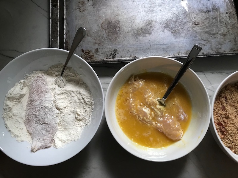 Raw chicken in bowl with flour and in a bowl with eggs for Oven Fried Chicken Breast. The chicken is coated with Bacon Crumbs and Honey and it's healthier because it's baked, not fried, but still gives you all of the crispy, crunchy goodness! It also gives you sweet and salty crunch with the addition of honey and bacon!