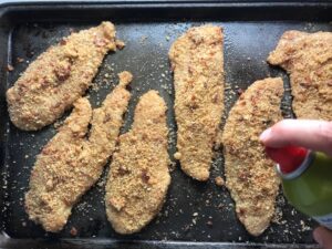 Hand spraying oil on raw chicken coated in bacon breadcrumbs on pan for Oven Fried Chicken Breast. The chicken is coated with Bacon Crumbs and Honey and it's healthier because it's baked, not fried, but still gives you all of the crispy, crunchy goodness! It also gives you sweet and salty crunch with the addition of honey and bacon!