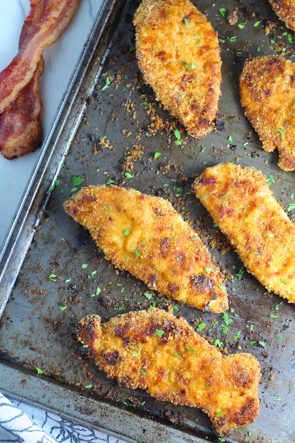Crispy Baked Chicken Breast Cutlets with Bacon Crumbs and Honey cooked on a sheet pan with parsley sprinkled on top and slices of bacon on the counter.