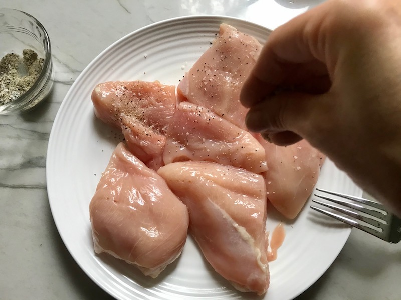 Hand seasoning raw chicken on plate for Creamy Bacon Chicken with Tomatoes. The chicken is smothered in a thickened sauce infused with smokey and salty bacon flavor. Cherry tomatoes give a sweet and tangy pop.
