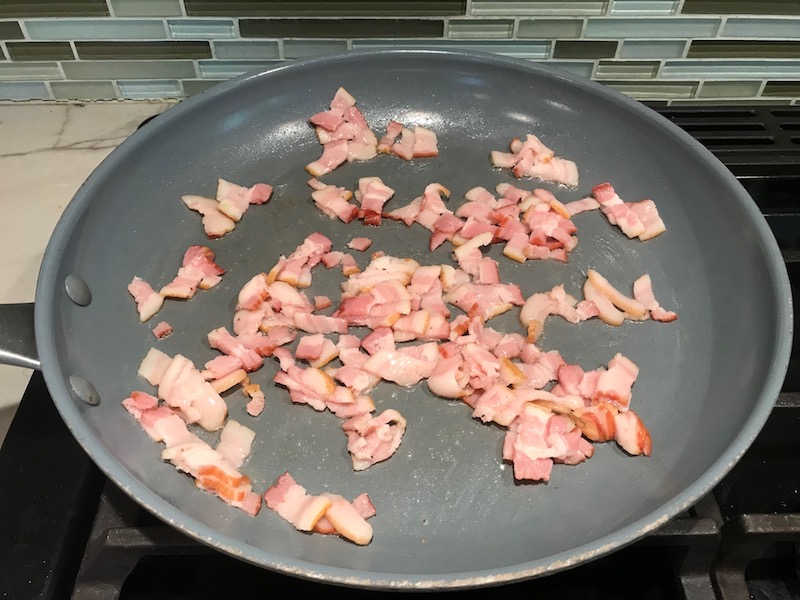 Bacon bits cooking in pan for Smothered Chicken with Bacon, and Tomatoes. The chicken is smothered in a thickened sauce infused with smokey and salty bacon flavor. Cherry tomatoes give a sweet and tangy pop.