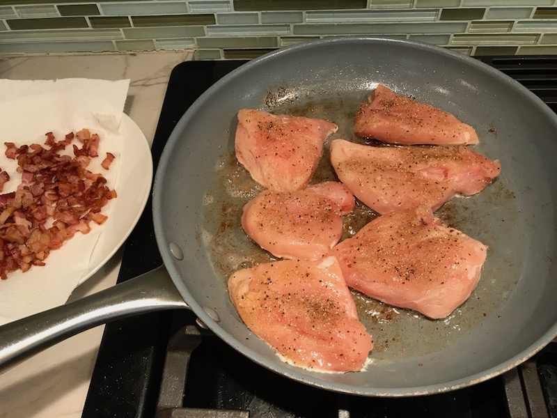 Chicken searing in pan with bacon bits on a plate to the side for Smothered Chicken with Bacon, and Tomatoes. The chicken is smothered in a thickened sauce infused with smokey and salty bacon flavor. Cherry tomatoes give a sweet and tangy pop.