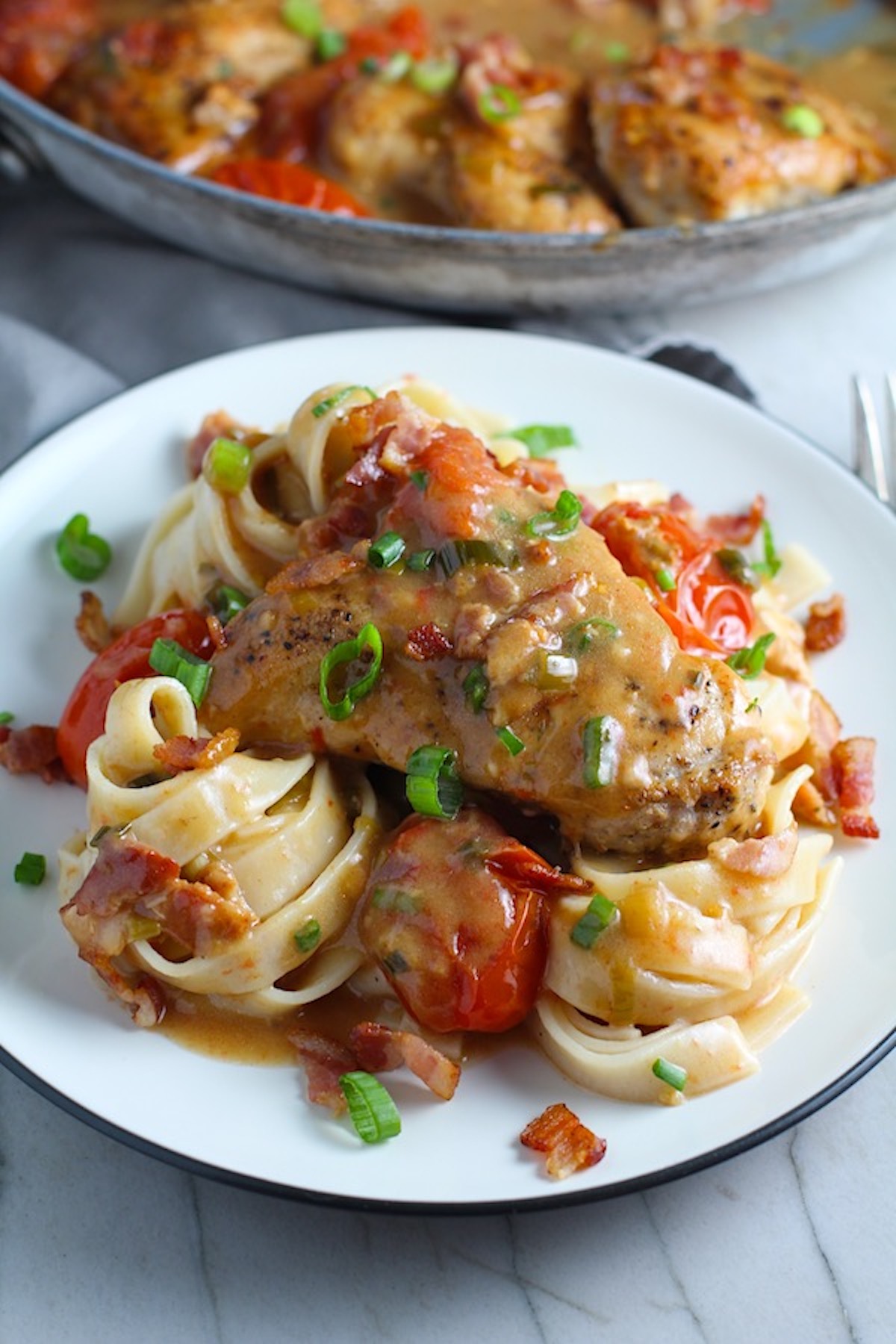 Creamy Bacon Chicken with Tomatoes and fettuccine on a plate with pan in background. The chicken is smothered in a thickened sauce infused with smokey and salty bacon flavor. Cherry tomatoes give a sweet and tangy pop.