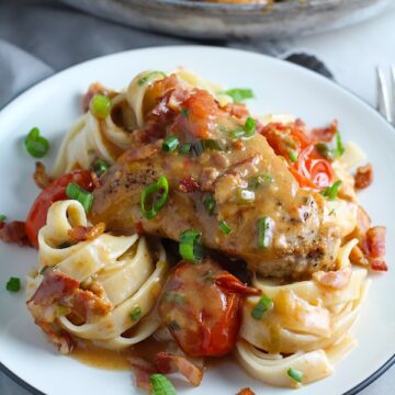 Smothered Chicken with Bacon, and Tomatoes and fettuccine on a plate with pan in background. The chicken is smothered in a thickened sauce infused with smokey and salty bacon flavor.  Cherry tomatoes give a sweet and tangy pop.