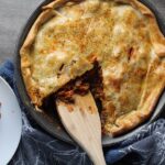 Savory Empanada Beef Pie Recipe in a skillet with spatula and piece out on counter. It has a perfect flaky, buttery pastry on the outside with a savory, smokey, salty ground beef filling.  The best part is that everything cooks in one skillet!