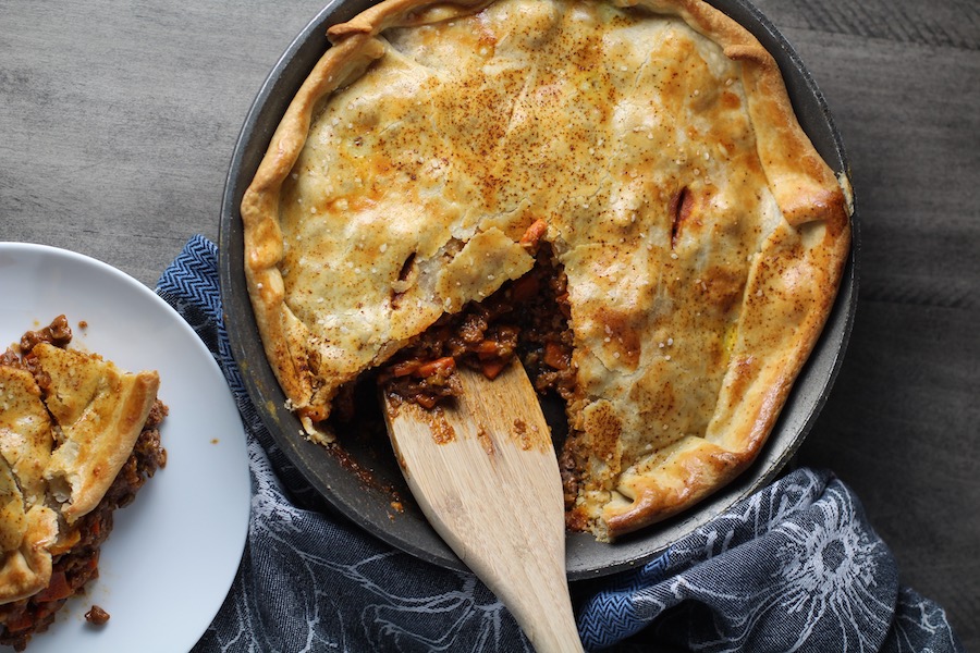 Flaky Empanada Pie with Ground Beef in a skillet with a piece out on a plate next to it and wooden spatula in the empty piece section in the skillet.