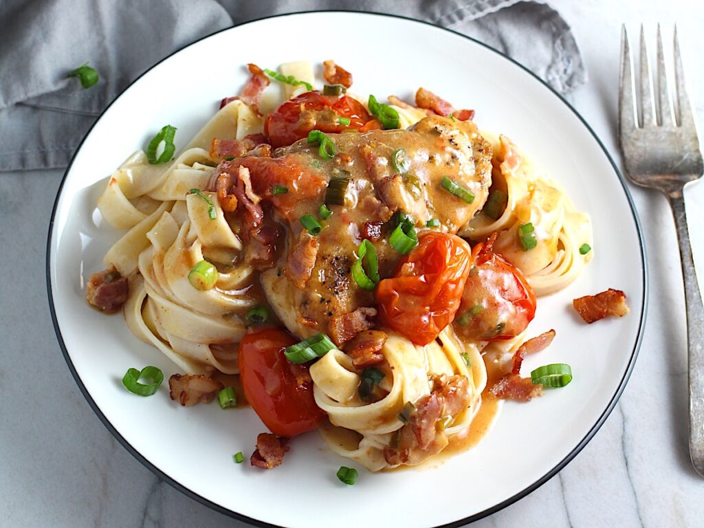 Smothered Chicken with Bacon, and Tomatoes and fettuccine on a plate with pan in background. The chicken is smothered in a thickened sauce infused with smokey and salty bacon flavor. Cherry tomatoes give a sweet and tangy pop.