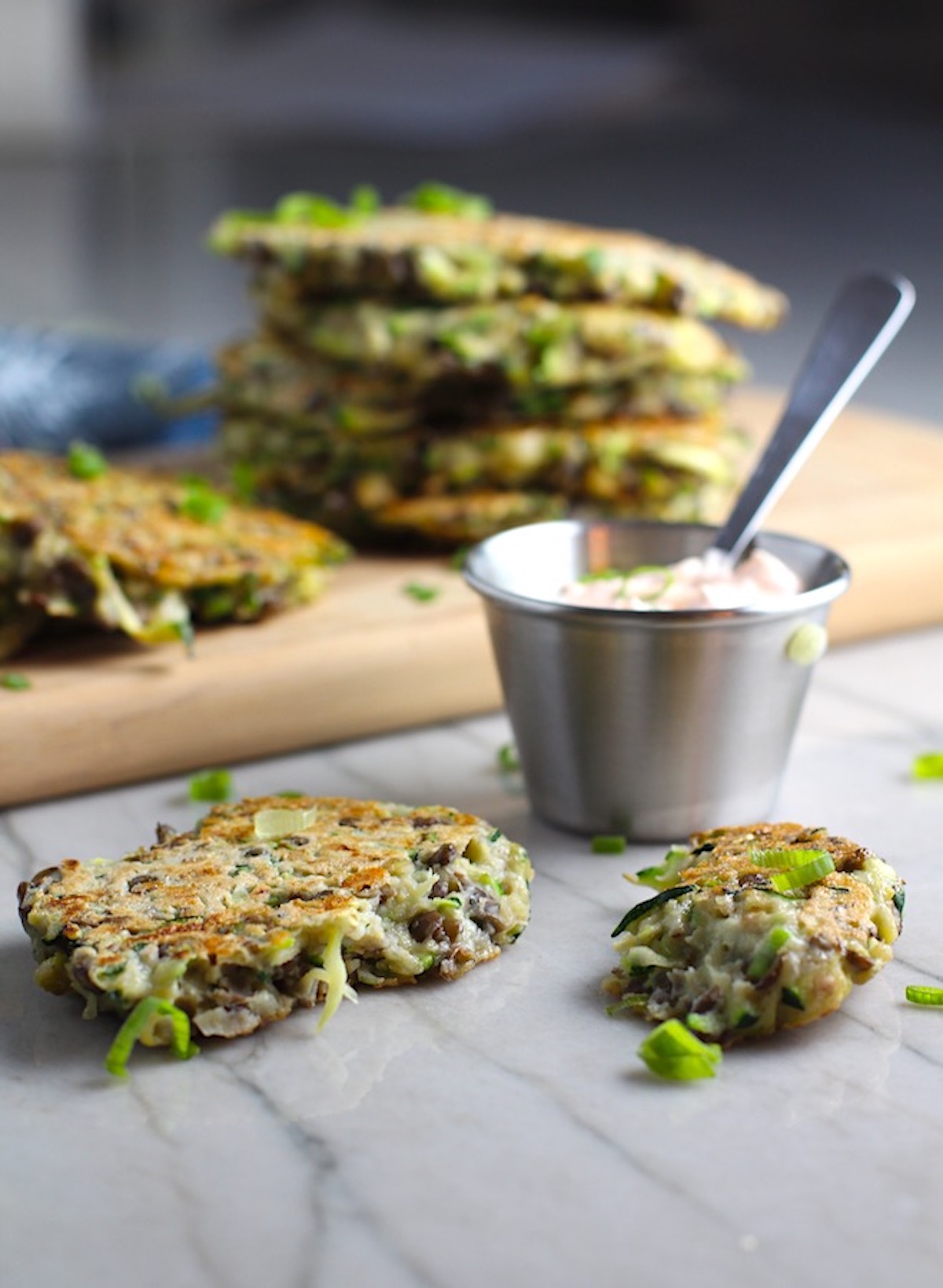 Zucchini Lentil Fritters broken in two on counter with more on a cutting board in the background and Sriracha sauce on the side. They have a crispy outside and soft inside and are perfect for an easy dinner! The flavors are simple but perfect with parmesan, oregano, scallions, lentils, and zucchini.