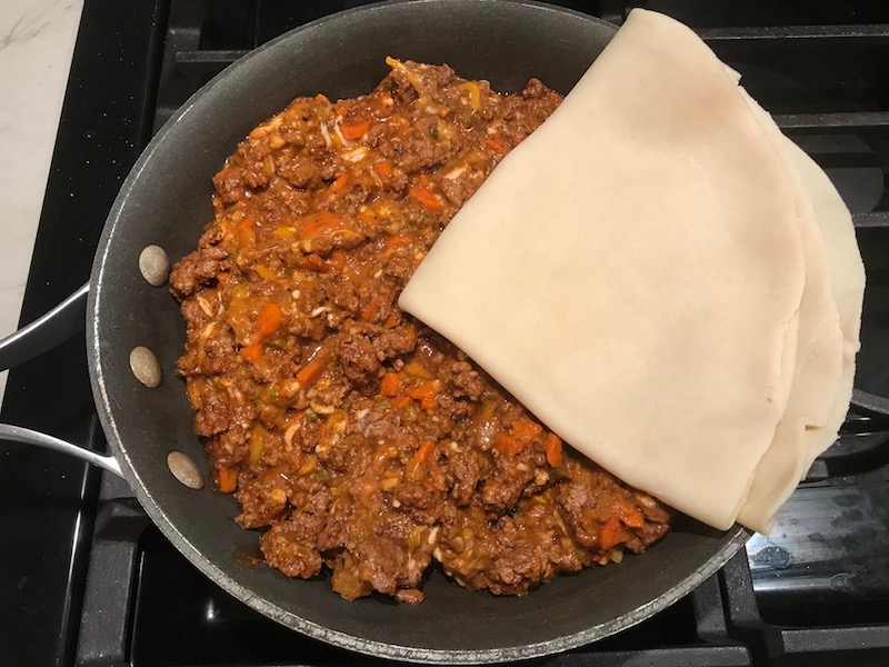 Beef mixture in skillet and pie crust being added to top for Empanada Beef Pie Recipe. It has a perfect flaky, buttery pastry on the outside with a savory, smokey, salty ground beef filling. The best part is that everything cooks in one skillet!