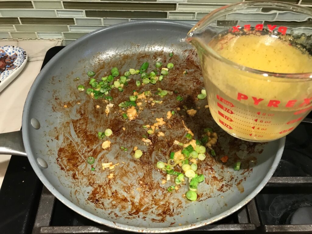 Broth about to be poured into a skillet with browned bits on the bottom and scallions and garlic for Creamy Paprika Chicken.