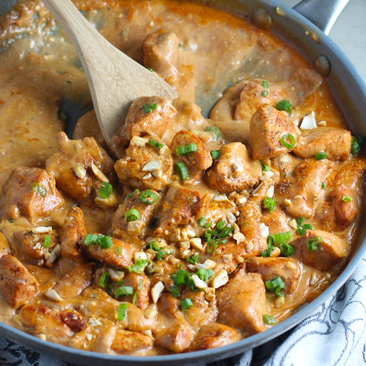 Quick Cooking Creamy Paprika Chicken in a frying pan with wood spoon and scallions and chopped cashews on top. It's creamy, smokey, rich, and hearty. This is an easy weeknight family dinner!