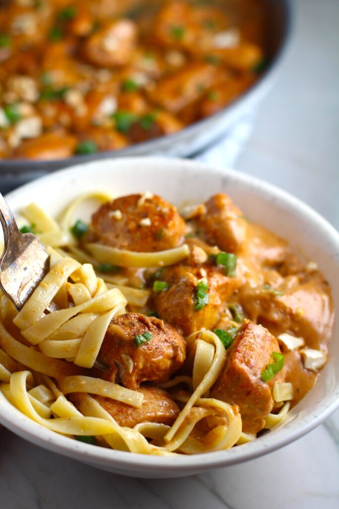 Quick Cooking Creamy Paprika Chicken in a bowl with pasta and fork scooping pasta and pan in background. It's creamy, smokey, rich, and hearty. This is an easy weeknight family dinner!