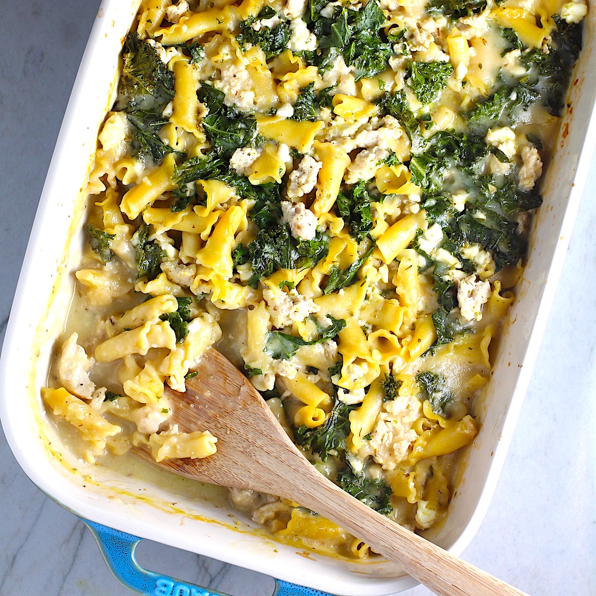 Greek Ground Chicken Feta Pasta Casserole in a casserole dish with wood spatula. It has lean ground chicken with garlic, oregano, parmesan, feta and Kale.