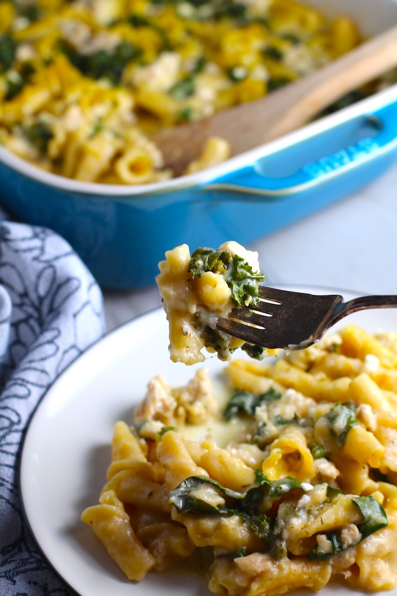 Fork with bite of Glorious Greek Ground Chicken Pasta Casserole with plate below and casserole in background. It has lean ground chicken with garlic, oregano, parmesan, feta and Kale.