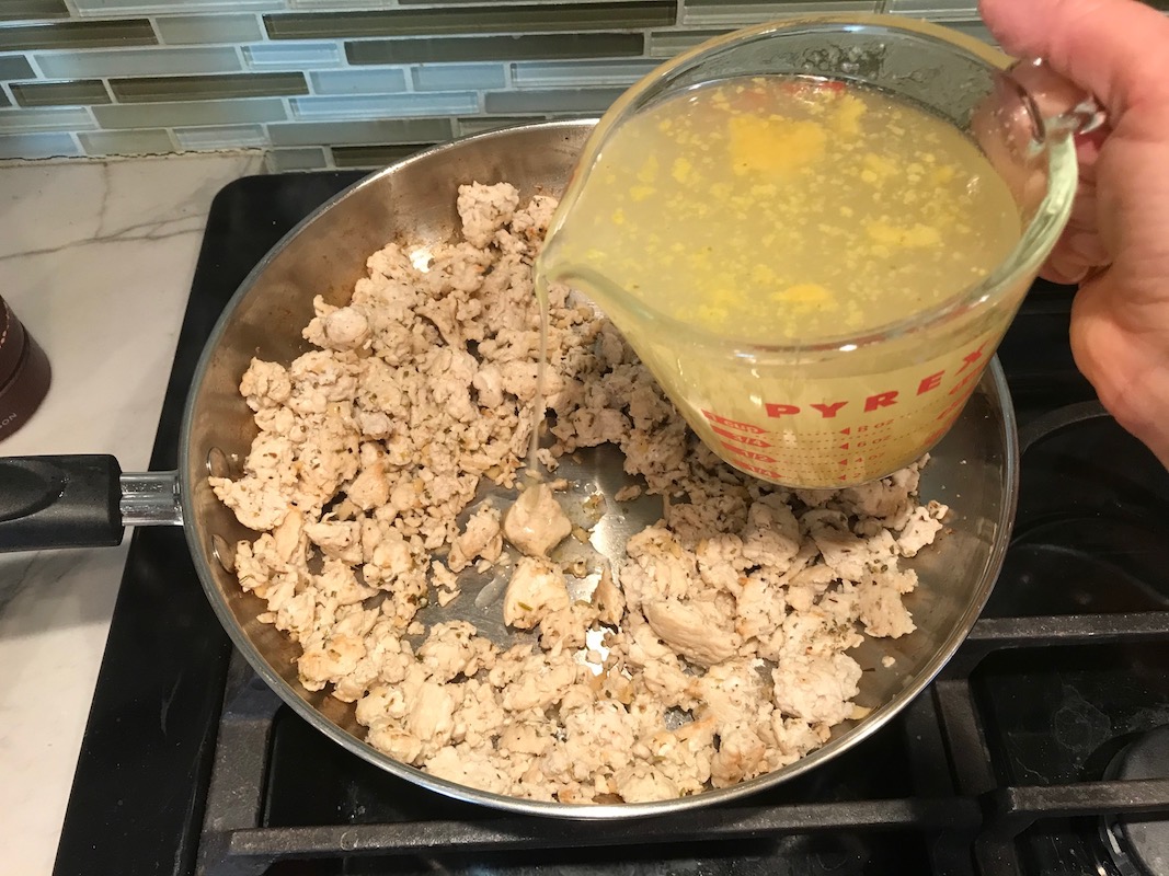 Measuring cup of broth being added to ground chicken in pan cooking for Greek Ground Chicken Feta Pasta Casserole. It has lean ground chicken with garlic, oregano, parmesan, feta and Kale.
