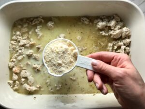 Hand adding measuring cup of parmesan cheese to dish with ground chicken and broth for Greek Ground Chicken Feta Pasta Casserole. It has lean ground chicken with garlic, oregano, parmesan, feta and Kale.