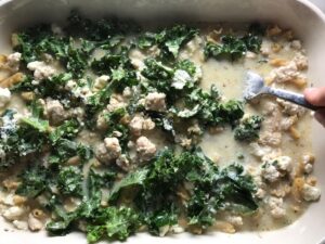 Fork pressing down dry pasta and kale into liquid with ground chicken and broth for Greek Ground Chicken Feta Pasta Casserole. It has lean ground chicken with garlic, oregano, parmesan, feta and Kale.