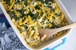 Greek Ground Chicken Feta Pasta Casserole in a casserole dish with wood spatula. It has lean ground chicken with garlic, oregano, parmesan, feta and Kale.