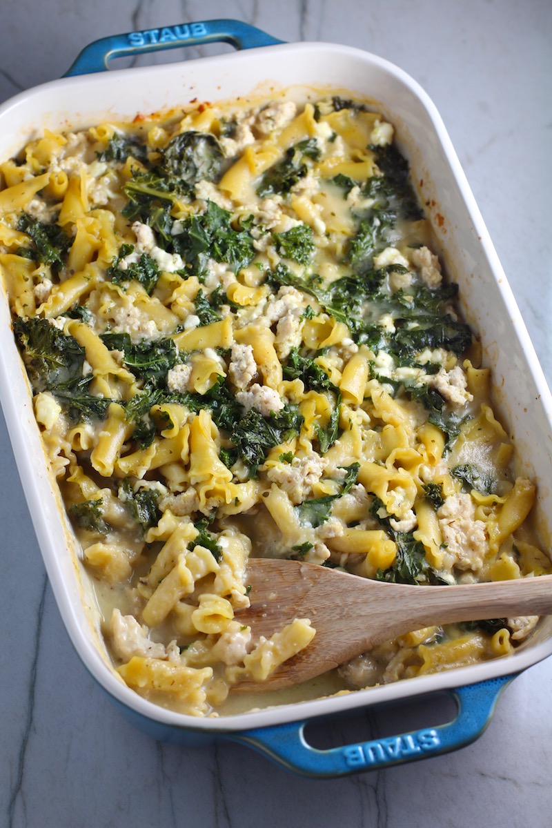 Greek Ground Chicken Feta Pasta Casserole in a casserole dish with wood spatula. It has lean ground chicken with garlic, oregano, parmesan, feta and Kale. 