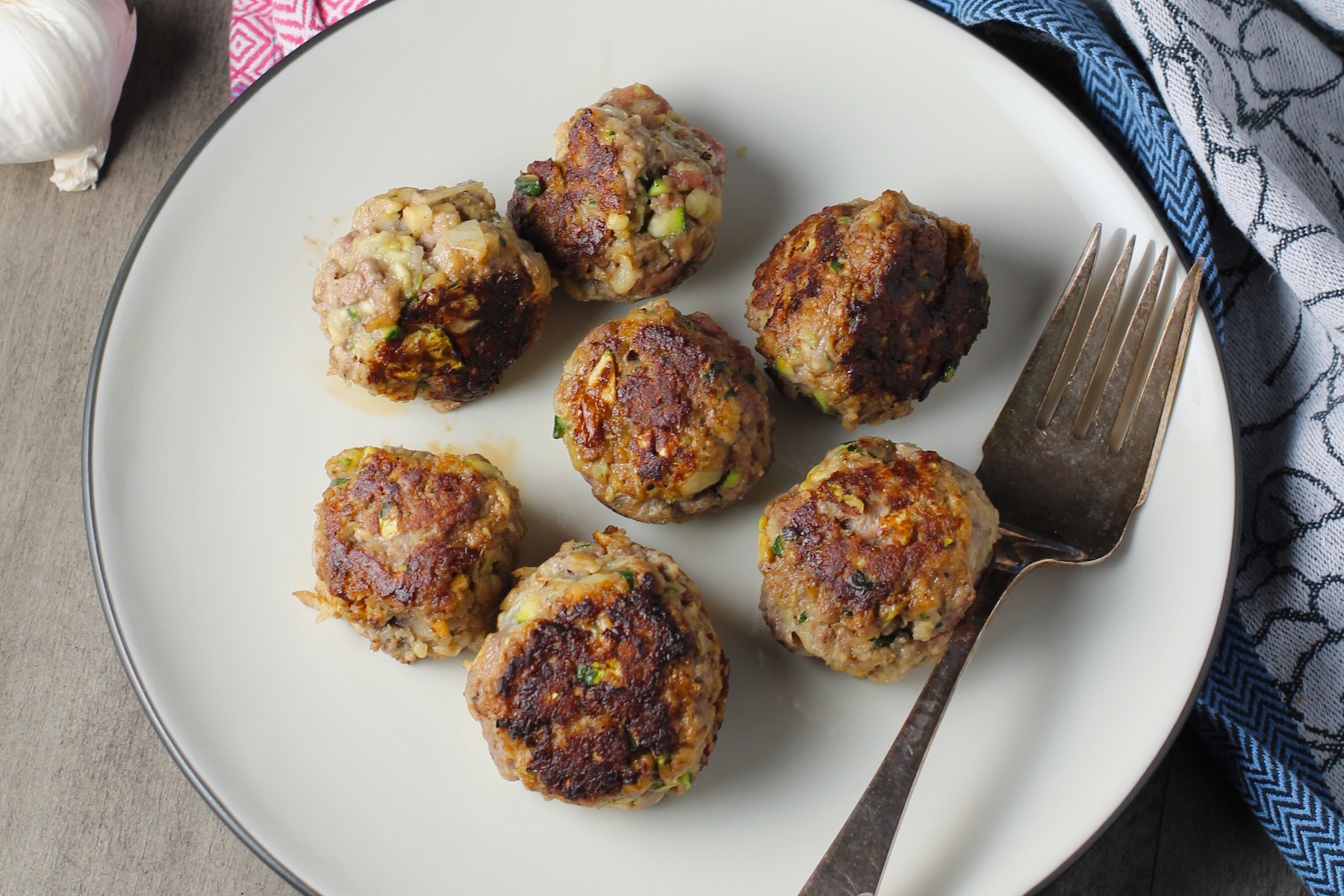 Healthy Meatballs with Zucchini & Lean Beef on a plate with fork and pan in background. They're loaded with flavor from the garlic, minced onion, salt, and pepper. The zucchini brings moisture, texture, and bulks up the meatballs with healthiness.