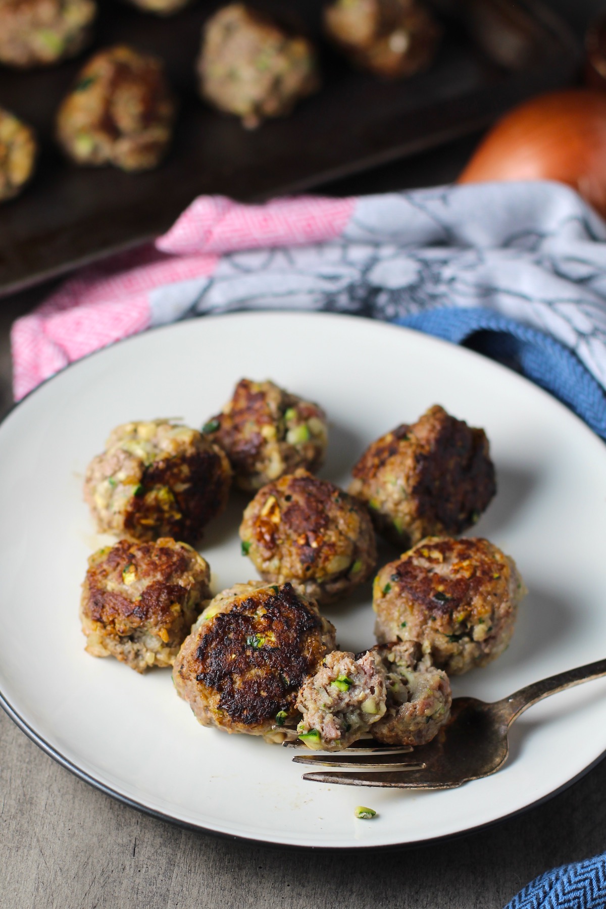 Healthy Meatballs with Zucchini & Lean Beef on a plate with fork cutting into one and pan in background.  They're loaded with flavor from the garlic, minced onion, salt, and pepper.  The zucchini brings moisture, texture, and bulks up the meatballs with healthiness.