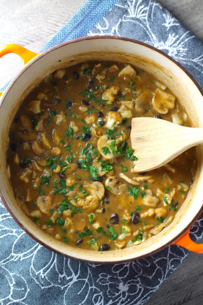 Creamy Cajun Cauliflower and Beans in apot with wood spoon. The mild cajun flavors combined with healthy garbanzo beans, black beans, cauliflower, and mushrooms make this a hearty, creamy, and delicious dish