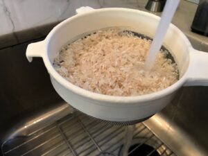 Rinsing uncooked rice under sink in strainer for Saffron Butter Crispy Rice. Saffron brings a floral note and beautiful yellow-orange color. Added to the butter is what gives this crispy rice the buttery, crispy bottom.