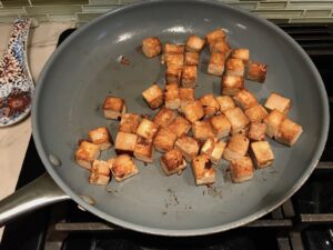 Firm teriyaki-marinated tofu cut into cubes searing in a hot pan for Thai Peanut Sauce Noodles with seared tofu and veggies.