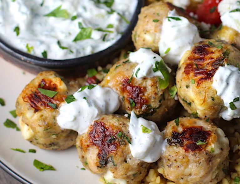 Ground Chicken Greek Meatballs on a plate with Tzatziki sauce over rice. Baked meatballs in back. These meatballs have garlic, feta, parsley, and oregano.