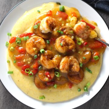 Cajun Shrimp and Grits recipe on a plate. It has seared shrimp in a creamy smoky cajun sauce with chicken andouille sausage and pimiento. Served over creamy, cheesy grits.