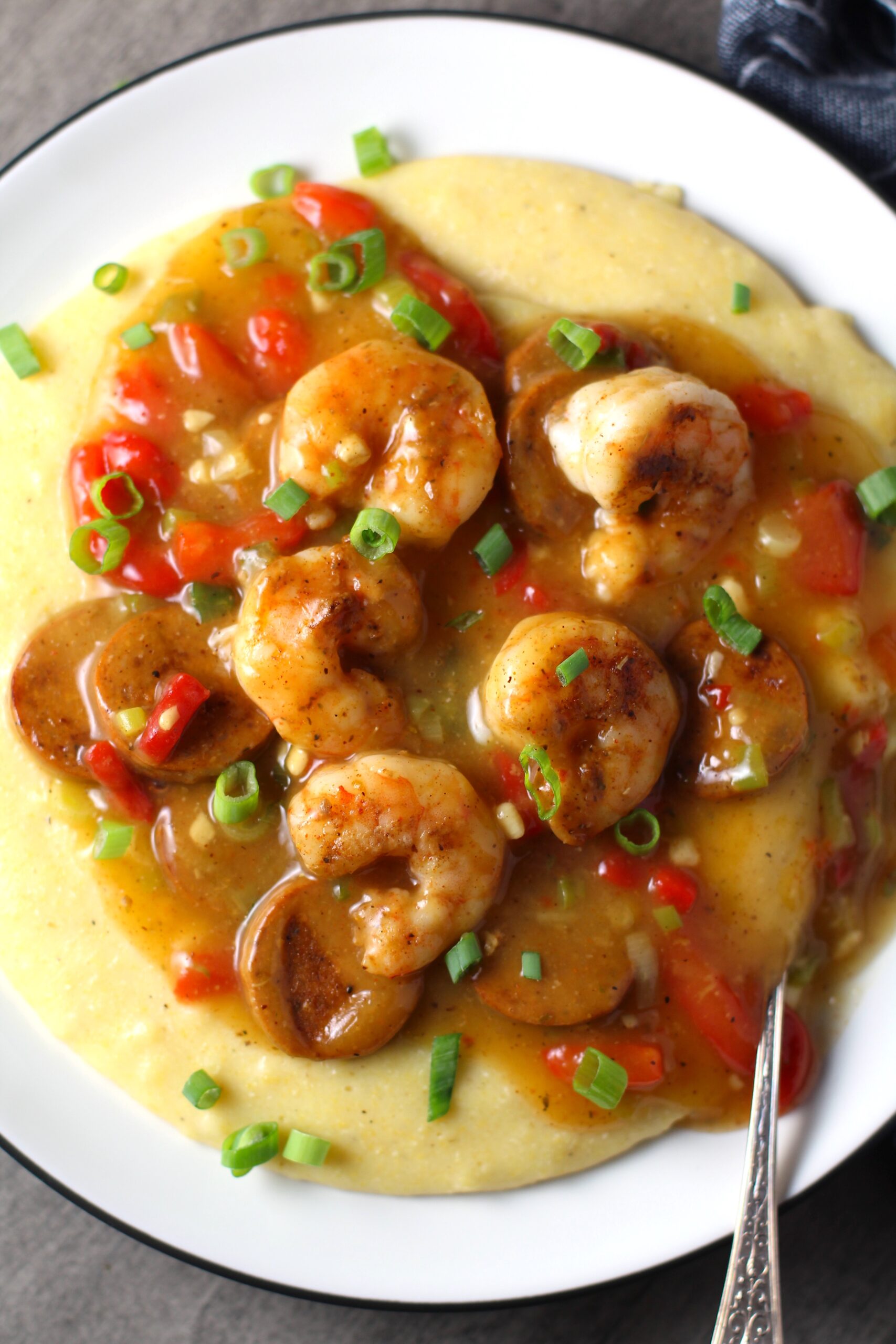 Cajun Shrimp and Grits recipe on a plate. It has seared shrimp in a creamy smoky cajun sauce with chicken andouille sausage and pimiento. Served over creamy, cheesy grits.