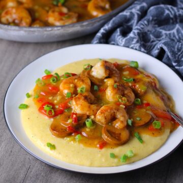 Cajun Shrimp and Grits recipe on a plate with pan in background. It has seared shrimp in a creamy smoky cajun sauce with chicken andouille sausage and pimiento.  Served over creamy, cheesy grits.