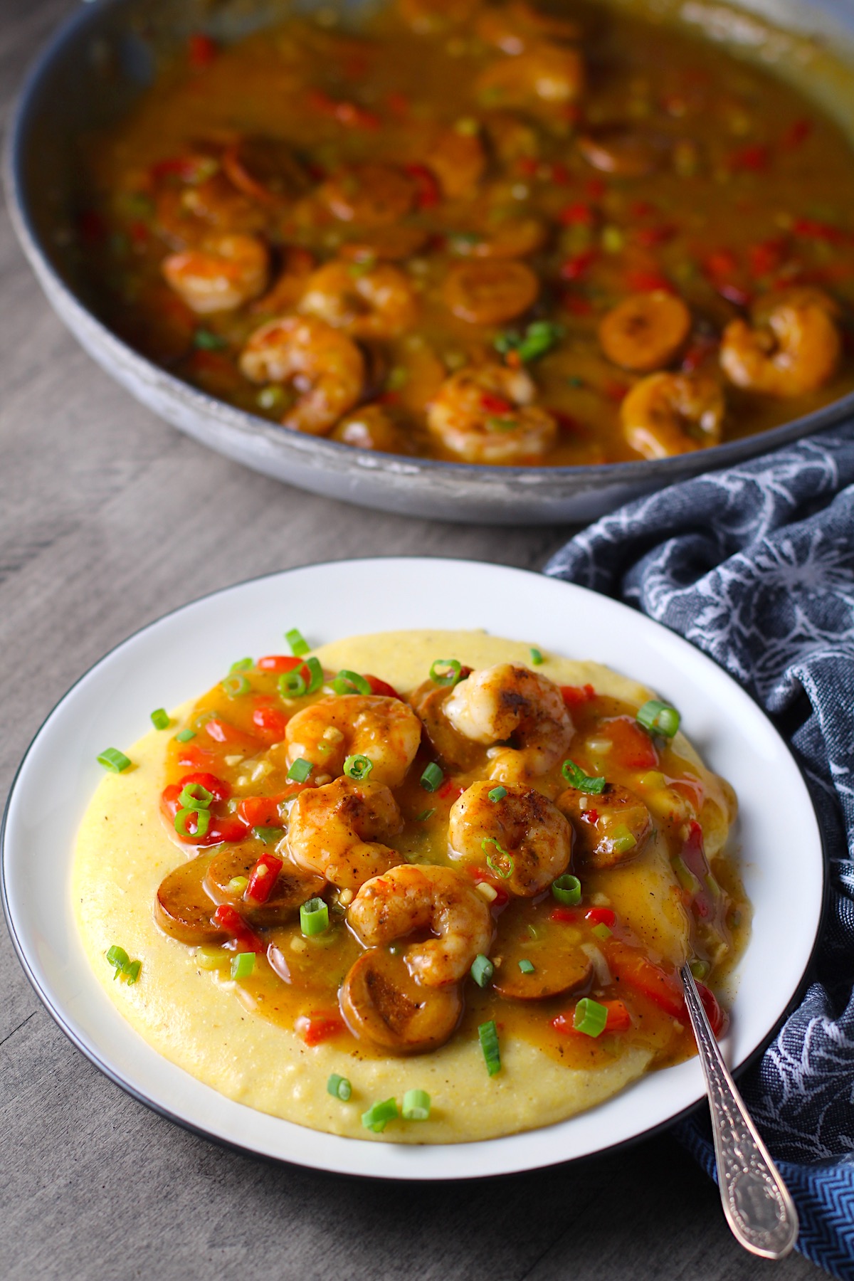 Cajun Shrimp and Grits recipe on a plate with pan in background. It has seared shrimp in a creamy smoky cajun sauce with chicken andouille sausage and pimiento. Served over creamy, cheesy grits.
